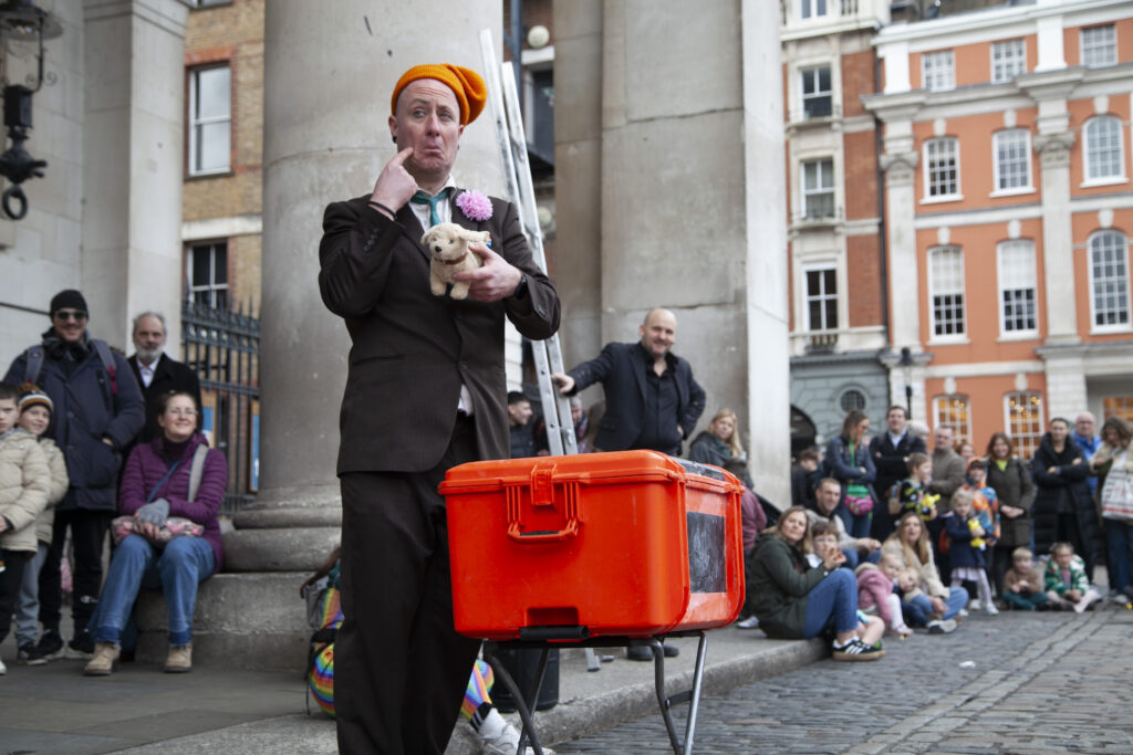 A man in a brown suit and an orange hat holds a stuffed toy dog looking sad